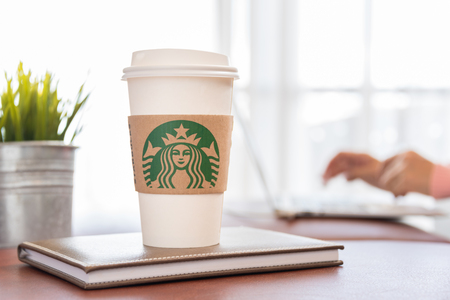 Bangkok, Thailand - October 5, 2016: Starbucks paper coffee cup on the table with woman sitting relaxing at home. Starbucks brand is one of the world famous from USA.のeditorial素材