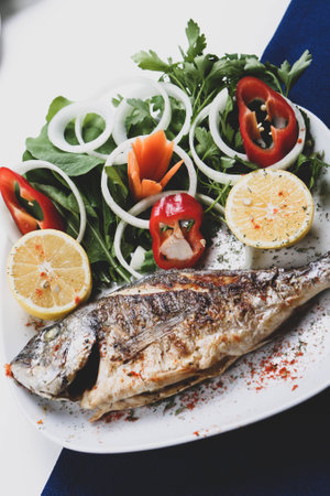 Fried sea bream (cupra) fish on white plate with onion, garden rocket and lemon slicesの写真素材