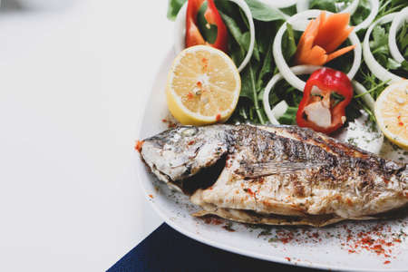 Fried sea bream (cupra) fish on white plate with onion, garden rocket and lemon slicesの写真素材