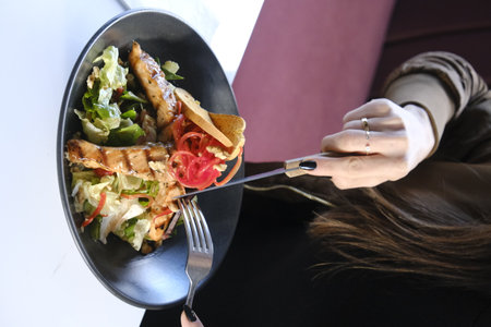 Woman eating salad with meat in restaurant, closeup of hands with fork and knifeの写真素材