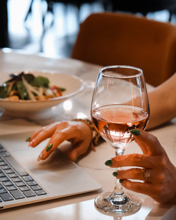 Close up of female hands holding glass of rose wine and working on laptop in cafeの写真素材