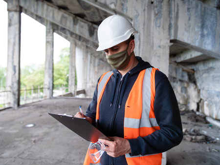 An engineer at a construction site in a construction helmet and medical mask records construction...