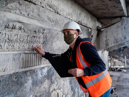 A male engineer in a construction helmet and antiviral mask inspects an emergency building for restoration or repair.の写真素材