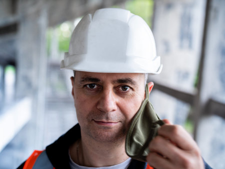 Portrait of a male Builder in a construction helmet and a medical antiviral mask. The man removes...