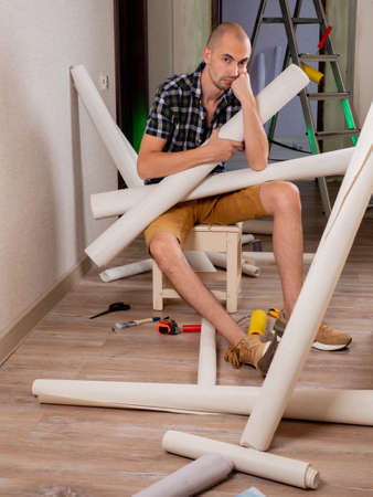 Renovation of the house. Preparation for wallpapering. A sad man sits on a bench and holds rolls of Wallpaper in his hands.の写真素材