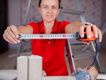 A woman holding a measure tape in her hands on the background of a stepladder and rolls of wallpaper.の写真素材