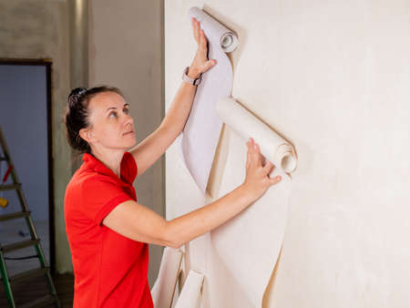 Woman in red shirt trying out new wallpaper at home holding the roll up against a newly painted white wall for visual effectの写真素材