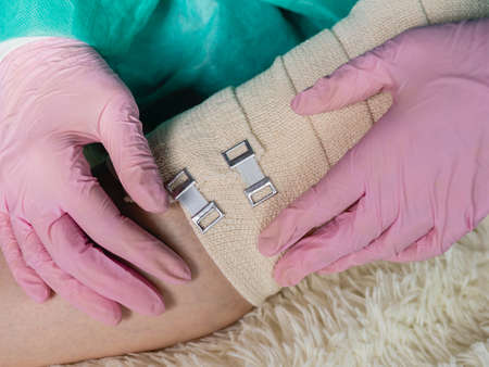 A doctor wearing medical gloves wraps an elastic bandage around the patients leg and knee. The traumatologist applies an elastic material to the patientの写真素材