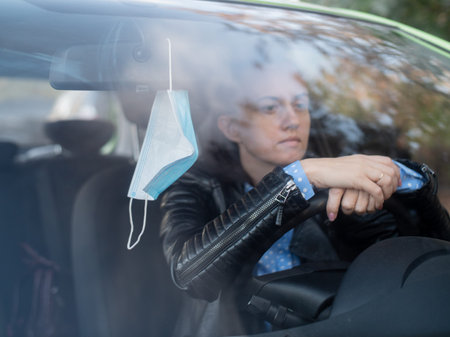 Sad woman driver sits in the car and holds his hands on the steering wheel of the car. Medical mask...