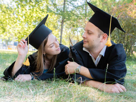 Happy loving couple of graduates in mantles lie on the lawn in the park of the universityの写真素材