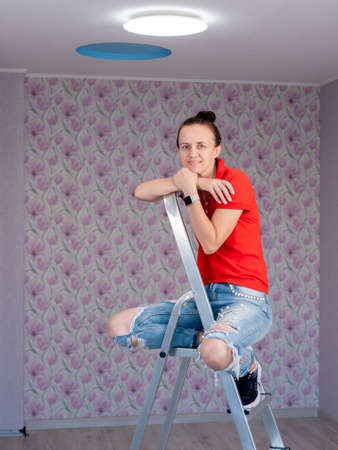 A contented woman is sitting on a stepladder in a room with newly pasted wallpaper and a new LED ceiling light. The repair is over.の写真素材