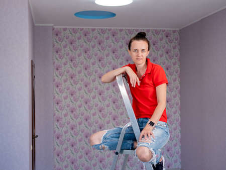 A contented woman is sitting on a stepladder in a room with newly pasted wallpaper and a new LED ceiling light. The repair is over.の写真素材