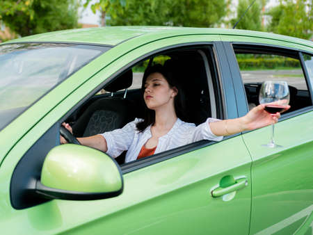 Young drunk girl driving a car with a glass of wine in her hand. Drinking alcohol while driving. a girl while driving a car stuck her hand out the window with a glass of wineの写真素材