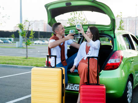 Beautiful young couple with glasses of wine in their hands stand next to the car and suitcases packed for summer travelの写真素材