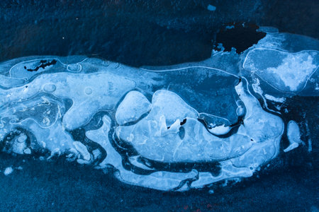 Ice with air bubbles and fancy fault lines, covered with snow in places , abstract pattern, background. Blue ice texture with dark water flowing under it. Frozen water on a lake in a winter forestの写真素材