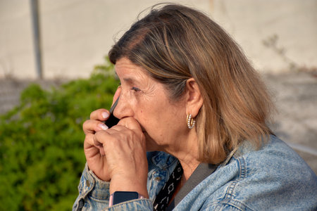 An elderly woman is seated, talking on her mobile phone with a serious expression on her face. Her...