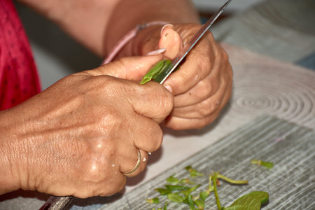 The hands of an elderly woman as she carefully slices green beans with a knife, preparing them for...