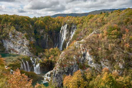 Autumn scene captures the enchanting waterfalls at Plitvice Lakes National Park in Croatia, surrounded by the vibrant colors of fall. The golden and orange hues of the leaves create a magical contrast against the cascading water, adding to the serene beauty of the park. The tranquil lakes reflect the vivid autumn colors, while the mist rising from the waterfalls gives the image a mystical atmosphere. Plitvice Lakes, a UNESCO World Heritage Site, is renowned for its pristine nature and captivating landscapes, especially during the fall season.の写真素材