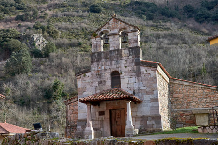 This image showcases the historic church of Pola de Somiedo, a charming village in Asturias, Spain. Nestled in a breathtaking valley surrounded by towering green mountains, this traditional Asturian church stands as a testament to the region's rich cultural and architectural heritage. The simple yet elegant stone facade, complemented by a classic bell tower, reflects the timeless beauty of northern Spain's rural churches. The surrounding landscape, with its lush greenery and rolling hills, enhances the peaceful and spiritual atmosphere of the scene. This church serves as a focal point for the local community, preserving traditions and history in a region known for its natural beauty and deep-rooted customs. Ideal for illustrating themes of history, religion, travel, and rural European architecture, this photograph captures the essence of a tranquil Spanish village.の写真素材