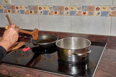 A close-up of a grandmother's hands holding wooden cooking utensils while preparing a meal on a modern stovetop. The image highlights the blend of tradition and modernity, capturing the warmth of home cooking. Ideal for themes related to family, culinary heritage, and lifestyle photography.の写真素材