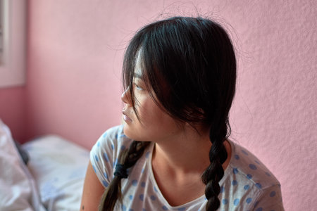 A young Chinese woman with autism in a moment of introspection, gazing off into the distance with a calm and thoughtful expression. Her hair is styled in pigtails, and she is seated against a soft pink wall in a serene indoor setting. This photograph was taken in commemoration of World Autism Awareness Day, celebrated annually on April 2, to promote understanding and acceptance of neurodiversity and the unique beauty of individuality.の写真素材