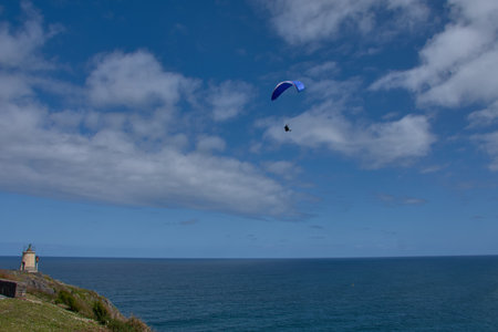 A paraglider enjoys a breathtaking flight above Faro de San Agustin in Coana, Asturias, Spain. Captured against a dramatic cloudy sky, the scene highlights the beauty of extreme sports and outdoor adventure in northern Spain. The surrounding wind turbines and mountainous landscape create a stunning contrast with the glider's vibrant canopy. Perfect for themes of freedom and exploration.の写真素材