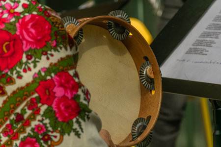 A detailed close-up of wrinkled, wise hands delicately holding and playing a wooden tambourine with pale heads and metal jingles. The colorful traditional costumes of Galician cantoeiras, adorned with flowers and embroidery, create a vibrant background evoking the cultural richness and joy of folk music, ideal for illustrating themes of tradition, music, Galician culture, festivals, and celebrations.の写真素材