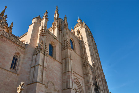 The Cathedral of Segovia stands tall with intricate gothic details, bathed in golden morning light under a vibrant blue sky. Its symmetrical composition, vertical lines, and warm tones convey grandeur and history, ideal for themes of architecture, religion, travel, and European heritageの写真素材