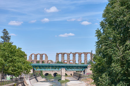The monumental stone and brick pillars of the Aqueduct of Miracles, a jewel of Roman engineering, rise above a blanket of green grass in the city of Merida. The image, with its vibrant color contrast (ochre, green, and blue) and the juxtaposition of the ancient ruin with the modern city in the background, is ideal for content about history, travel, archaeology, and World Heritage.の写真素材