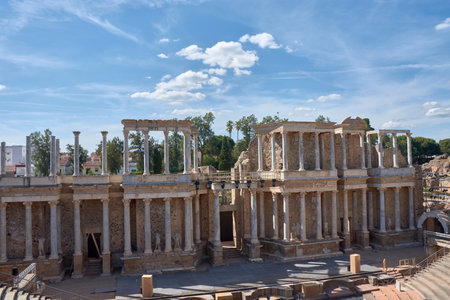 The imposing archaeological remains of the Roman theater in Merida are presented with their majestic faÃ§ade of columns and sculptures under a partly cloudy sky. The composition highlights the grandeur and detail of this ancient structure, evoking the rich history and cultural legacy of the Roman Empire, ideal for projects on archaeology, history, cultural tourism, classical art, or ancient performance venues.の写真素材