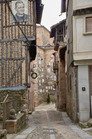 an evocative cobblestone alley in the village of Mogarraz, Salamanca, flanked by traditional houses with wooden balconies and stone walls. A distinctive element is the photographic portraits of residents adorning the facades, a project that pays homage to the town's identity and history. The linear composition guides the eye through the street, revealing the unique fusion of architectural heritage and cultural expression, ideal for publications on rural tourism, urban art, local history, or the authenticity of Spanish villagesの写真素材