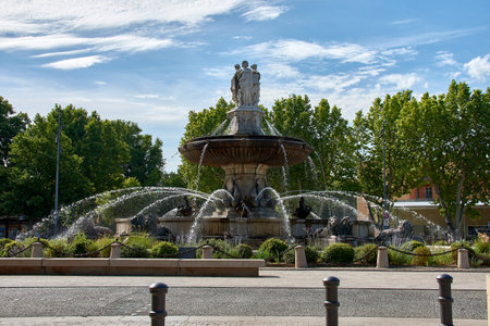 Monumental stone fountain with classical sculptures and multiple water jets, located in a tree-lined square in Aix en Provence. The blue sky with scattered clouds and sunlight provide a cheerful, summery atmosphere. Ideal image for editorial, tourism, or promotional purposes for cultural and heritage destinations.の写真素材