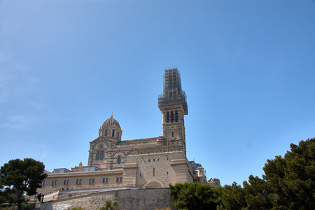 Notre Dame de la Garde, the iconic Neo-Byzantine-style basilica of Marseille, appears with its main tower covered in scaffolding during a restoration phase. The contrast between the two-tone stone and the metal structure of the scaffolding, under a deep blue sky, highlights the historical importance and conservation efforts of this religious and tourist symbol. Ideal for projects on heritage, architecture, restoration, and cultural tourism in the South of France.の写真素材