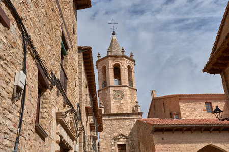 The tower of the Church of the Assumption in Cantavieja rises above a group of stone buildings that retain their medieval essence. The warm tones of the walls and ceilings contrast with the partly cloudy sky, creating a historic and authentic atmosphere. This photograph conveys a strong sense of cultural heritage and is ideal for editorial, tourism, or promotional projects about architecture and travelの写真素材