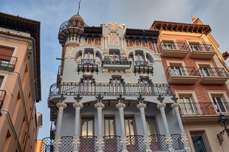 The El Torico Textile House in Teruel stands out with its modernist facade in soft tones and wrought iron balconies, surrounded by historic buildings. The warm light of the sunset highlights the ornamental details and the decorative dome, conveying a cultural and urban atmosphere. Ideal for publishing, tourism, and architectural projectsの写真素材