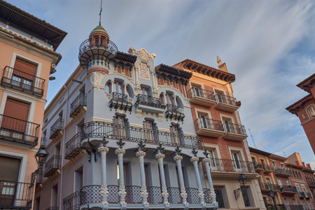The El Torico Textile House in Teruel stands out with its modernist facade in soft tones and wrought iron balconies, surrounded by historic buildings. The warm light of the sunset highlights the ornamental details and the decorative dome, conveying a cultural and urban atmosphere. Ideal for publishing, tourism, and architectural projectsの写真素材