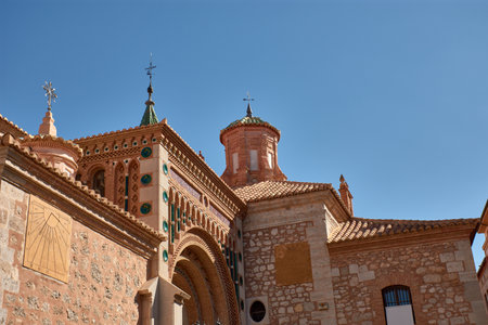 The tower of the Cathedral of Santa Maria de Mediavilla in Teruel rises in the Mudejar style with red brick, ornamental windows, and a green dome crowned by a metal cross. The blue sky highlights the texture of the materials and conveys serenity. Ideal for editorial, tourism, and cultural projects related to historical heritage and religious architectureの写真素材