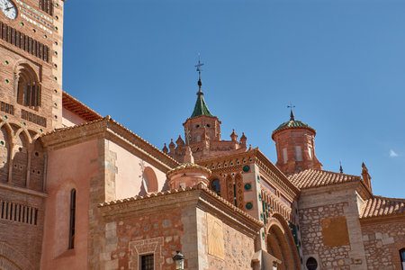 The tower of the Cathedral of Santa Maria de Mediavilla in Teruel rises in the Mudejar style with red brick, ornamental windows, and a green dome crowned by a metal cross. The blue sky highlights the texture of the materials and conveys serenity. Ideal for editorial, tourism, and cultural projects related to historical heritage and religious architectureの写真素材