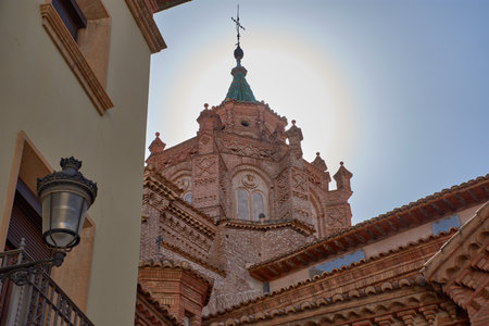 The tower of the Cathedral of Santa Maria de Mediavilla in Teruel rises in the Mudejar style with red brick, ornamental windows, and a green dome crowned by a metal cross. The blue sky highlights the texture of the materials and conveys serenity. Ideal for editorial, tourism, and cultural projects related to historical heritage and religious architectureの写真素材
