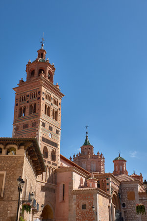 The tower of the Cathedral of Santa Maria de Mediavilla in Teruel rises in the Mudejar style with red brick, ornamental windows, and a green dome crowned by a metal cross. The blue sky highlights the texture of the materials and conveys serenity. Ideal for editorial, tourism, and cultural projects related to historical heritage and religious architectureの写真素材