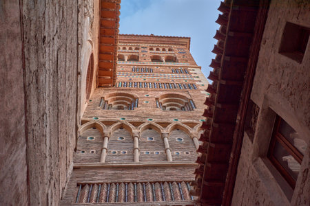 The imposing Mudejar Tower of San Martin, a recognized World Heritage Site, rises majestically with its distinctive reddish brick construction, elaborate geometric patterns of latticework and green and white tiles, and multiple arches and windows. Flanked by buildings with light-colored or stone facades, and framed by a sky alternating between intense blue and soft clouds, the tower exudes a sense of history and grandeur. The composition, which often plays with perspective from the narrow streets of Teruel, highlights the architectural, historical, and cultural richness of this iconic site, ideal for illustrating topics such as cultural heritage, Mudejar architecture, tourism in Spain, art history, travel to Teruel, and urban life with a historical touch.の写真素材