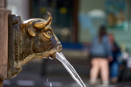 A richly patinated bronze sculpture representing a bull's head emerges from the wall, releasing a clear, thin stream of water that symbolizes the founding legend of Teruel and its rich cultural heritage in Aragon. The foreground composition, dominated by golden brown and green tones of metal against a dark, abstract background, emphasizes the historic texture and dynamic action of water.の写真素材