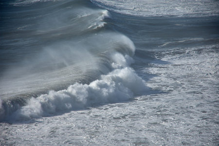 From a cliff, several surfers can be seen challenging the giant waves of Nazare, Portugal, in a high-energy marine environment. The contrast between the deep blue ocean, the white foam, and the moving human figures creates a powerful and dynamic composition. Ideal for depicting extreme sports, active tourism, and coastal landscapes with a high visual impactの写真素材