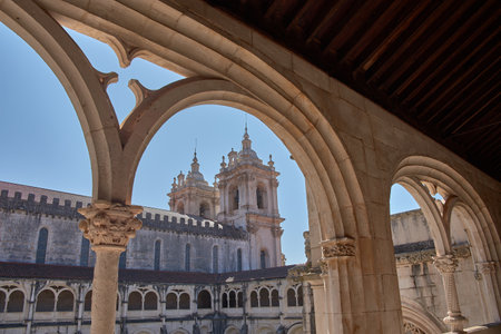 From an elevated gallery of the Alcobaca Monastery, the main facade is framed by carved stone arches, creating a symmetrical and profound composition. The blue sky and natural light enhance the architectural details and bring serenity to the environment. Ideal for representing historical heritage, religious architecture, and cultural tourism in Portugalの写真素材