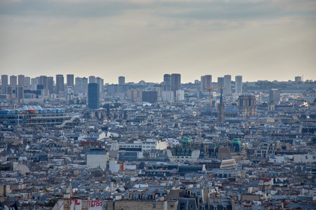 From the hill of Montmartre, a panoramic view of Paris unfolds, revealing gray rooftops, narrow streets, and an urban fabric that stretches to the horizon. The partly cloudy sky adds depth and serenity, while the surroundings evoke the bohemian and artistic spirit of the neighborhood. Ideal for tourism, editorial, and cultural content about urban heritage, aerial photography, and Parisian lifeの写真素材