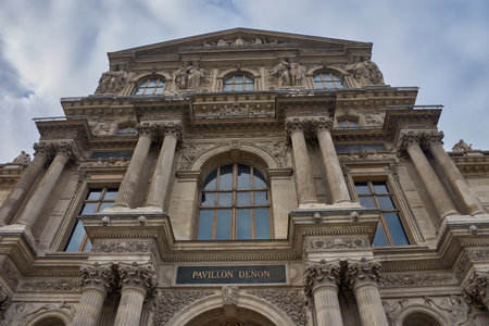 The facade of the Louvre Museum commands attention with its classical architecture, marked by columns, sculptures, and arches that evoke its past as a royal palace. The cloudy sky adds drama to the scene, while the symmetry and ornamental detail reinforces the historical character of the complex. Ideal for tourism, editorial, and cultural content on architectural heritage, museology, and European artの写真素材