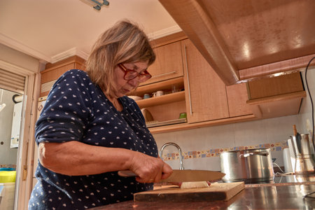 A 71-year-old woman prepares a vegetarian meal in her pajamas, chopping vegetables on a wooden board in a home kitchen. The warm lighting, vibrant colors of the ingredients, and everyday setting reinforce the authenticity of the scene, evoking healthy habits, active aging, and real life in familiar spaces. Ideal for editorial, social, and cultural content about mindful eating, household routines, and traditional cooking.の写真素材