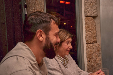 A 71-year-old woman smiles during a family dinner at a restaurant, accompanied by her children and her granddaughter of Chinese origin. The windows covered in raindrops from the nighttime rain add warmth and realism to the scene, conveying affection, celebration, and intergenerational connection. Ideal for editorial, social, and cultural content about family, active aging, and shared moments in urban settingsの写真素材