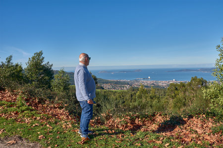 An elderly man leans on a wooden railing as he contemplates the Vigo Estuary from Monte Alba on a sunny day. The blue sky, clear light, and coastal landscape with buildings and mountains in the background reinforce the atmosphere of serenity and connection with the environment. Ideal for editorial content on local tourism, active aging, emotional well-being, and Galician natural heritage.の写真素材