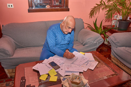 An elderly man is sitting in his living room, surrounded by bills, envelopes, and documents, he reviews them with concentration. The warm atmosphere, with pink walls, wooden furniture, and a decorative plant, conveys an authentic and emotional domestic scene. Ideal for campaigns on active aging, personal financial management, daily life, and independence in later life.の写真素材